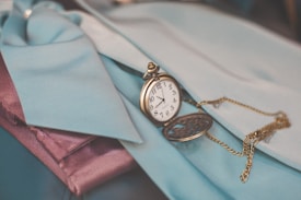 A vintage pocket watch with an ornate design rests on fabric in shades of teal and purple. The watch chain is intricately looped and creates a charming contrast with the surrounding materials.