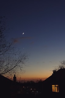 A crescent moon and a bright star are visible in a darkening sky during twilight. Silhouettes of houses and leafless trees frame the scene, with one house window emitting a warm glow.