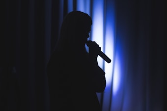 A silhouetted figure is holding a microphone against a backdrop of vertical blue-lit curtains, creating a dramatic and contemplative ambiance.