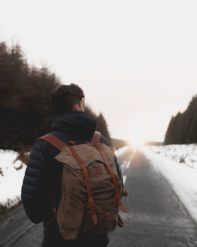 A person wearing a large backpack, standing on a snowy road lined with trees, facing towards a bright horizon. The scene suggests a journey or adventure taking place in a cold, tranquil environment.