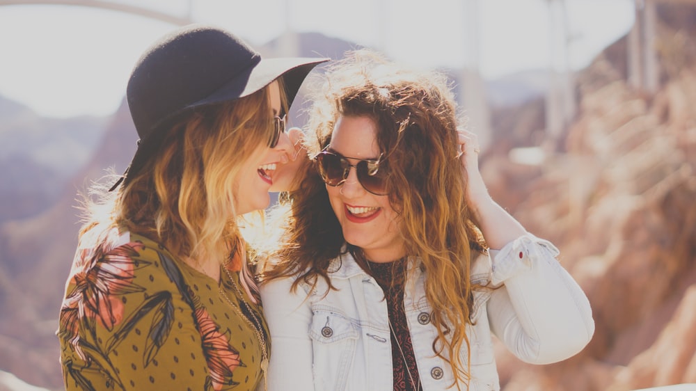 Two fashionable women at the Hoover Dam Access Road stand closely, laughing in the sunshine