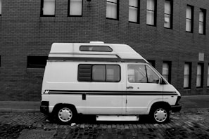 A vintage camper van is parked on a cobblestone street alongside a brick building. The van has a retro design with a stripe along the side and a raised roof. Several windows with Venetian blinds are visible on the building's facade.