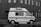 A vintage camper van is parked on a cobblestone street alongside a brick building. The van has a retro design with a stripe along the side and a raised roof. Several windows with Venetian blinds are visible on the building's facade.