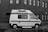 A vintage camper van is parked on a cobblestone street alongside a brick building. The van has a retro design with a stripe along the side and a raised roof. Several windows with Venetian blinds are visible on the building's facade.