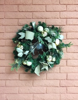 A decorative wreath adorned with green pine needles, white and gold ornaments, and beige ribbons hangs on a brick wall. The wreath features a blend of natural and festive elements, creating a holiday atmosphere.