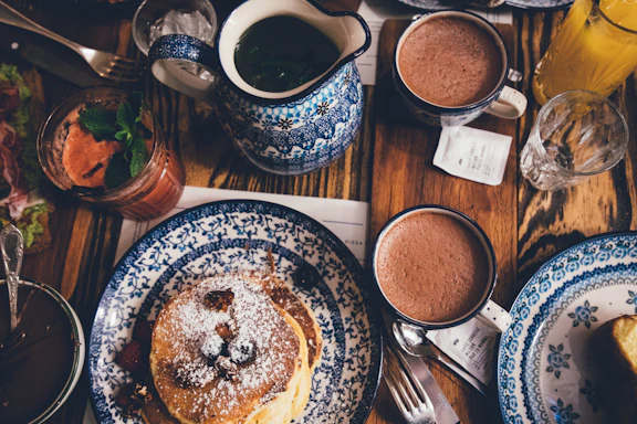 A warm, inviting table setting featuring soft beige linens, a ceramic cup of herbal latte, and a plate of oat pancakes with cream syrup, all bathed in natural light.