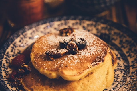 A stack of fluffy pancakes topped with powdered sugar, blueberries, and walnuts is served on a decorative blue and white plate. Raspberries and additional nuts are scattered around the pancakes, creating a warm and inviting presentation.