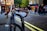 A parked bicycle with blue handlebars is locked to a black bike rack on a cobblestone street. The background shows a busy urban scene with people walking and buildings featuring shopfronts and a red telephone booth.