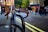 A parked bicycle with blue handlebars is locked to a black bike rack on a cobblestone street. The background shows a busy urban scene with people walking and buildings featuring shopfronts and a red telephone booth.
