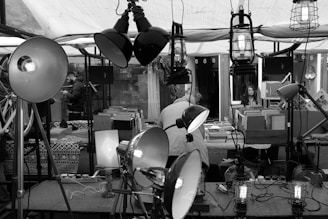 A busy market stall features an array of lamps and light fixtures displayed prominently. In the background, a woman is seated at a counter covered with books and boxes, suggesting a focus on selling second-hand items. Various objects, including bicycles and patterned textiles, are visible. The setting conveys an eclectic and vintage market vibe.
