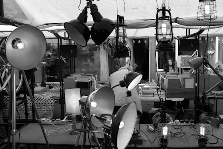 A busy market stall features an array of lamps and light fixtures displayed prominently. In the background, a woman is seated at a counter covered with books and boxes, suggesting a focus on selling second-hand items. Various objects, including bicycles and patterned textiles, are visible. The setting conveys an eclectic and vintage market vibe.