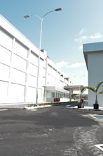 A modern industrial or commercial building with a clean, white facade. The scene includes a well-paved, empty road leading up to the main entrance. Tall street lights line the road, and there are neatly planted palm trees in planters adding a touch of greenery. The sky is clear and blue, indicating sunny weather.