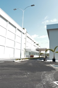 A modern industrial or commercial building with a clean, white facade. The scene includes a well-paved, empty road leading up to the main entrance. Tall street lights line the road, and there are neatly planted palm trees in planters adding a touch of greenery. The sky is clear and blue, indicating sunny weather.