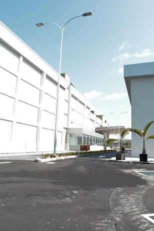A modern industrial or commercial building with a clean, white facade. The scene includes a well-paved, empty road leading up to the main entrance. Tall street lights line the road, and there are neatly planted palm trees in planters adding a touch of greenery. The sky is clear and blue, indicating sunny weather.