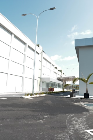 A modern industrial or commercial building with a clean, white facade. The scene includes a well-paved, empty road leading up to the main entrance. Tall street lights line the road, and there are neatly planted palm trees in planters adding a touch of greenery. The sky is clear and blue, indicating sunny weather.