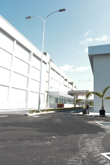 A modern industrial or commercial building with a clean, white facade. The scene includes a well-paved, empty road leading up to the main entrance. Tall street lights line the road, and there are neatly planted palm trees in planters adding a touch of greenery. The sky is clear and blue, indicating sunny weather.