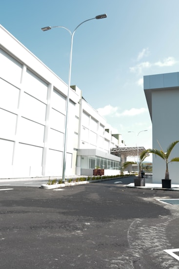 A modern industrial or commercial building with a clean, white facade. The scene includes a well-paved, empty road leading up to the main entrance. Tall street lights line the road, and there are neatly planted palm trees in planters adding a touch of greenery. The sky is clear and blue, indicating sunny weather.