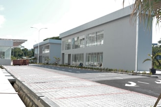 A modern industrial complex with two large, rectangular, gray buildings featuring large windows. The buildings are surrounded by a paved area with parking spaces and a few small palm trees. A large dumpster is placed on the paved surface near the buildings. Bright sunlight highlights the scene with clear skies above.