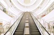A modern, multi-level shopping mall interior featuring a central escalator that directs the eye upward. The architecture showcases glossy floors, large glass railings, and a well-lit environment with plants and various store displays visible in the background.
