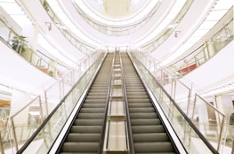 A modern, multi-level shopping mall interior featuring a central escalator that directs the eye upward. The architecture showcases glossy floors, large glass railings, and a well-lit environment with plants and various store displays visible in the background.