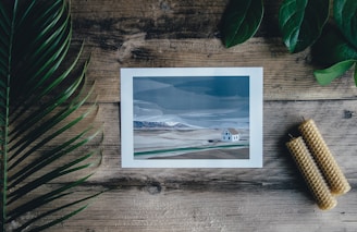 A printed image of a minimalist landscape with a house is placed on a wooden table. Surrounding the image are green leaves to the left and top right, and two rolled beeswax candles to the bottom right. The colors in the artwork are muted blues and grays, with shades of green and brown in the landscape.