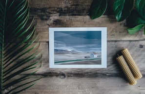 A printed image of a minimalist landscape with a house is placed on a wooden table. Surrounding the image are green leaves to the left and top right, and two rolled beeswax candles to the bottom right. The colors in the artwork are muted blues and grays, with shades of green and brown in the landscape.