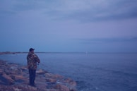 A person is standing on a rocky shore holding a fishing rod, casting into calm waters under a somewhat cloudy sky. The scenery appears serene and tranquil, with the horizon extending indefinitely.