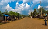 A rural street lined with simple buildings and lush green trees beneath a bright blue sky with fluffy white clouds. The road is unpaved and stretches into the distance, with only a few people walking. On the right, a person carries a large crate balanced on their head.