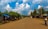 A rural street lined with simple buildings and lush green trees beneath a bright blue sky with fluffy white clouds. The road is unpaved and stretches into the distance, with only a few people walking. On the right, a person carries a large crate balanced on their head.