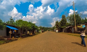 A rural street lined with simple buildings and lush green trees beneath a bright blue sky with fluffy white clouds. The road is unpaved and stretches into the distance, with only a few people walking. On the right, a person carries a large crate balanced on their head.