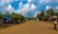 A rural street lined with simple buildings and lush green trees beneath a bright blue sky with fluffy white clouds. The road is unpaved and stretches into the distance, with only a few people walking. On the right, a person carries a large crate balanced on their head.
