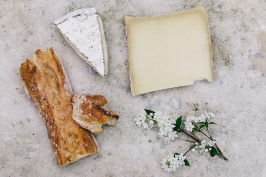 A rustic setting features two pieces of cheese, a broken piece of baguette, and small white flowers on a textured surface. One cheese piece is triangular with a white rind, and the other is rectangular with a pale yellow color. The baguette appears crusty and golden brown, and the flowers add a delicate touch.