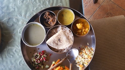 A traditional Indian thali is served on a round metal plate. It contains rice, a flatbread, several small bowls of various curries and lentils, along with a mix of vegetables and a spoon.