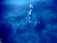 A person wearing flippers is diving into a deep blue underwater scene surrounded by small fish. The underwater landscape features large rock formations and varying shades of blue, creating a serene and adventurous atmosphere.