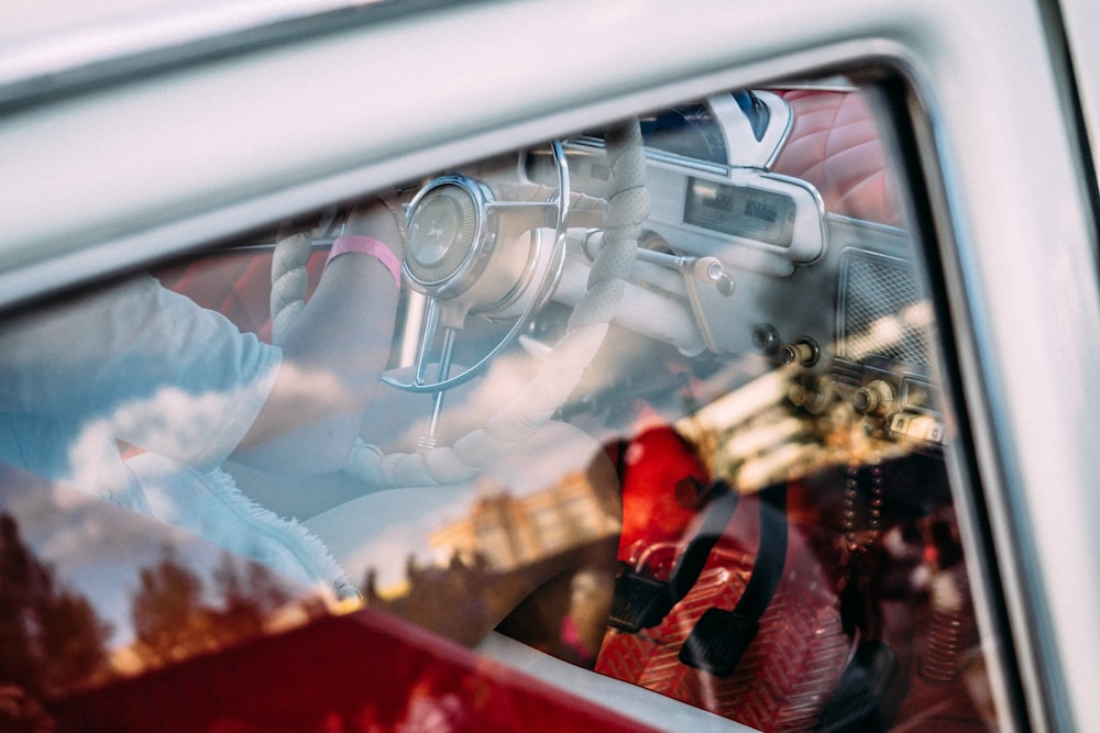 Partial view of a driver in a vintage car through the rear right window, overlapping with the reflection on the glass.