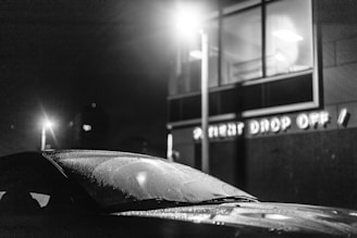 A black and white image featuring a car with a windshield covered in water droplets, parked near a building with illuminated windows. The wall of the building has signage that reads 'PATIENT DROP OFF'. A streetlight casts a glow, adding to the scene's nighttime ambiance.