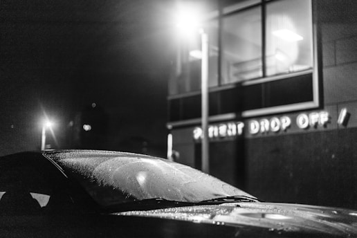 A black and white image featuring a car with a windshield covered in water droplets, parked near a building with illuminated windows. The wall of the building has signage that reads 'PATIENT DROP OFF'. A streetlight casts a glow, adding to the scene's nighttime ambiance.