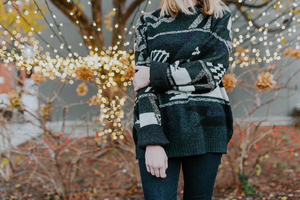 A woman wearing a cozy knit sweater standing in front of fairy lights and trees