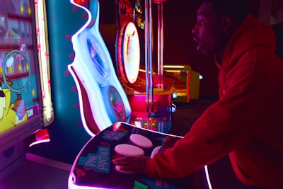 A person is intensely playing a brightly lit arcade game. The scene includes vibrant neon lights and a variety of gaming machines. The person is wearing a red hoodie and appears focused on the gameplay.
