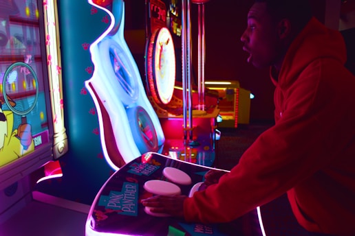 A person is intensely playing a brightly lit arcade game. The scene includes vibrant neon lights and a variety of gaming machines. The person is wearing a red hoodie and appears focused on the gameplay.