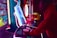 A person is intensely playing a brightly lit arcade game. The scene includes vibrant neon lights and a variety of gaming machines. The person is wearing a red hoodie and appears focused on the gameplay.