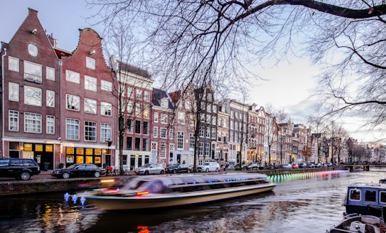 A canal runs through a cityscape with picturesque, traditional Dutch architecture. The buildings have classic gabled facades reflecting the historic style, and a boat moves swiftly through the water, creating a blurred effect. Vehicles are parked along the street, and bare trees frame the scene, indicating a late autumn or winter setting.