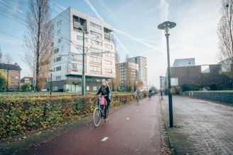 A street in an urban area with a dedicated bike lane, featuring several cyclists riding bicycles. The background shows modern apartment buildings and leafless trees, indicating a chilly but sunny day. Lamp posts line the path, and the scene conveys a sense of daily commuting or leisure cycling.