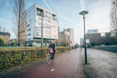 A street in an urban area with a dedicated bike lane, featuring several cyclists riding bicycles. The background shows modern apartment buildings and leafless trees, indicating a chilly but sunny day. Lamp posts line the path, and the scene conveys a sense of daily commuting or leisure cycling.
