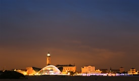A cityscape photograph taken during twilight, with buildings illuminated by artificial lights. The skyline includes a prominent tower that stands out among lower structures, and the sky transitions from a deep blue at the top to a warm orange hue near the horizon.
