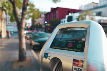 A close-up view of a parking meter with the option to insert coins or use a credit card. The background features a city street lined with parked cars and buildings, some of which are painted in vibrant colors like red. There are trees along the sidewalk.