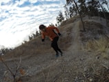 A person in an orange jacket and black pants is running uphill on a rocky trail surrounded by a natural landscape with sparse trees. The sky is partially cloudy and the terrain appears rugged with some dry grass and small bushes.