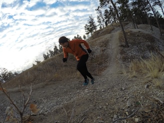 A person in an orange jacket and black pants is running uphill on a rocky trail surrounded by a natural landscape with sparse trees. The sky is partially cloudy and the terrain appears rugged with some dry grass and small bushes.