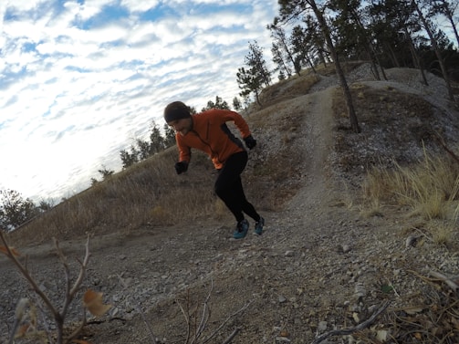 A person in an orange jacket and black pants is running uphill on a rocky trail surrounded by a natural landscape with sparse trees. The sky is partially cloudy and the terrain appears rugged with some dry grass and small bushes.