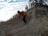 A person in an orange jacket and black pants is running uphill on a rocky trail surrounded by a natural landscape with sparse trees. The sky is partially cloudy and the terrain appears rugged with some dry grass and small bushes.